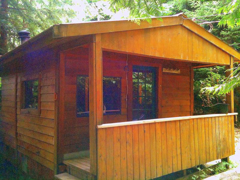 A small wooden cabin with a covered porch standing in a sunlit forest clearing